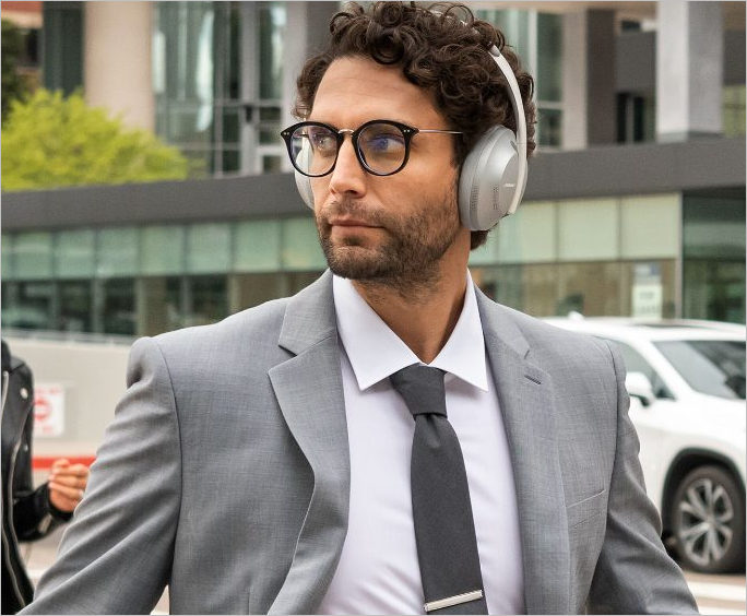 A man in a gray suit, looking to his right, wearing the silver variant of the Bose Noise Cancelling Headphones 700 outdoors