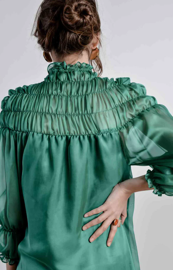  Close up of the back of the woman wearing a green smocked turtleneck. She rests her right hand on the small of her back.
