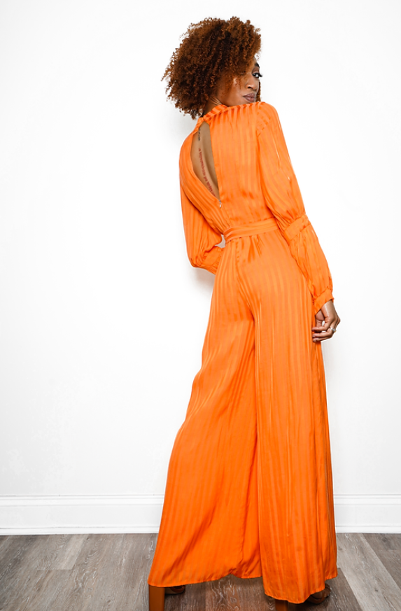 : A woman in an orange jumpsuit with long sleeves and a high neckline standing on wooden floor is seen from the back.