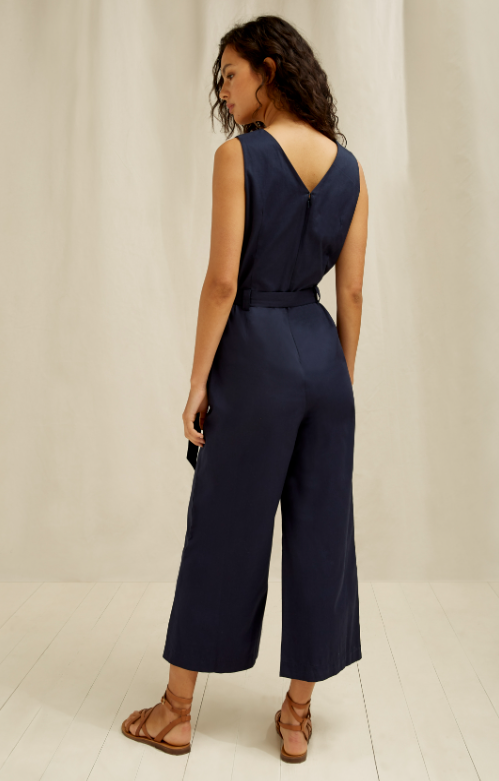 Woman wearing a sleeveless navy jumpsuit and a pair of flat strappy brown sandals is seen from the back.