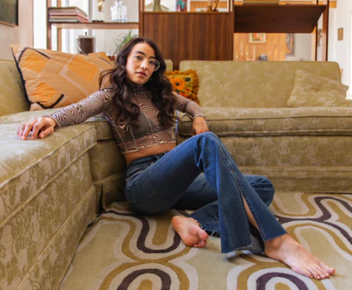 Woman in a long-sleeved sheer crop top and a pair of flared pants sitting on the floor against a moss green couch.