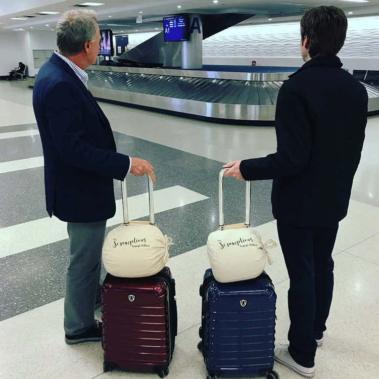 Two men standing beside their luggage with a travel pillow attached at an airport.