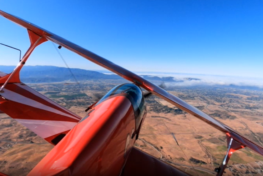 A red biplane soaring over a valley with majestic mountains in the background. Captured by GoPro HERO12 Black.