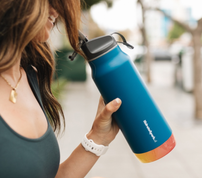 A woman holding a blue smart water bottle with a yellowish-red glow at the bottom of the bottle, the HidrateSpark PRO. Stay hydrated in style!